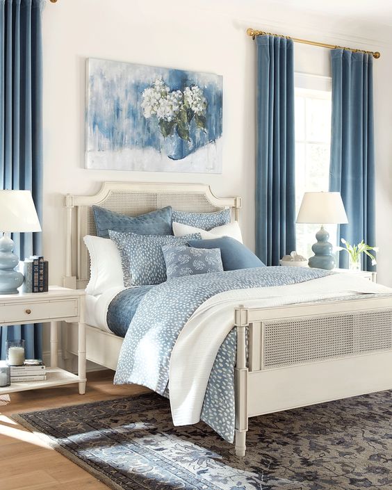 a stylish blue and white bedroom with a cane bed and white nightstands, blue bedding, a printed rug, blue curtains and an artwork