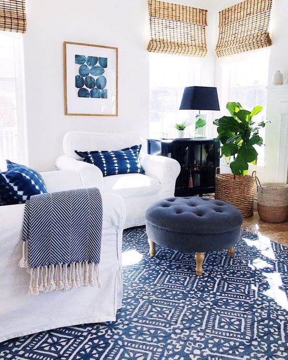 a stylish living room with white chairs, navy pillows, a navy pouf, a blue printed rug, a black sideboard, a black table lamp and woven shades