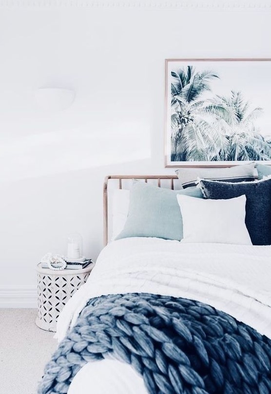 A tropical beach bedroom with a tropical artwork, a wooden bed, coastal colored textiles, a white perforated nightstand