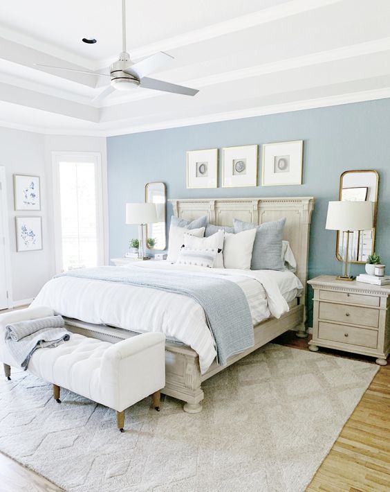 a welcoming bedroom with a pastel accent wall, a bed with blue and white bedding, a white upholstered bench, stained nightstands