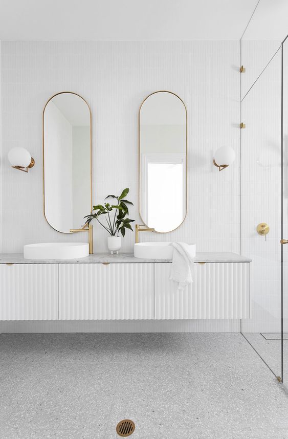 a white bathroom with a grey terrazzo floor, a shower space, a white fluted vanity with a stone countertop, vessel sinks and oval mirrors