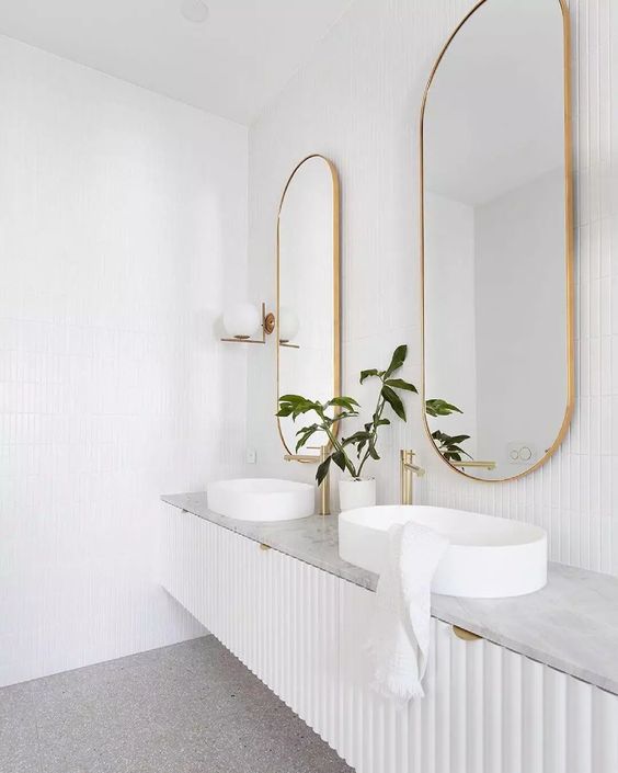 an airy bathroom with a white reeded vanity and a stone countertop, vessel sinks, gold fixtures and oval mirrors in gold frames