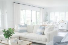 an airy coastal living room with a white and pale blue sofa, printed pillows, a wooden folding table, greenery and a neutral printed rug