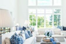 an airy living room with a double-height ceiling, neutral sofas, blue pillows, poufs and an ottoman, blue blooms