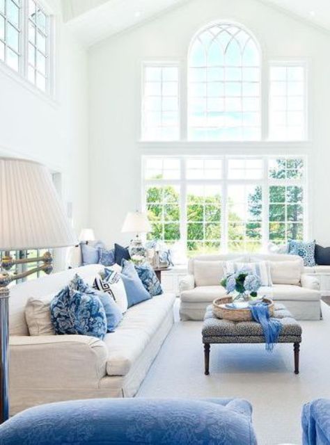 An airy living room with a double height ceiling, neutral sofas, blue pillows, poufs and an ottoman, blue blooms