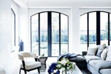 an airy living room with arched windows with black framing, a blue rug, grey blue sofas and white chairs, a coffee table