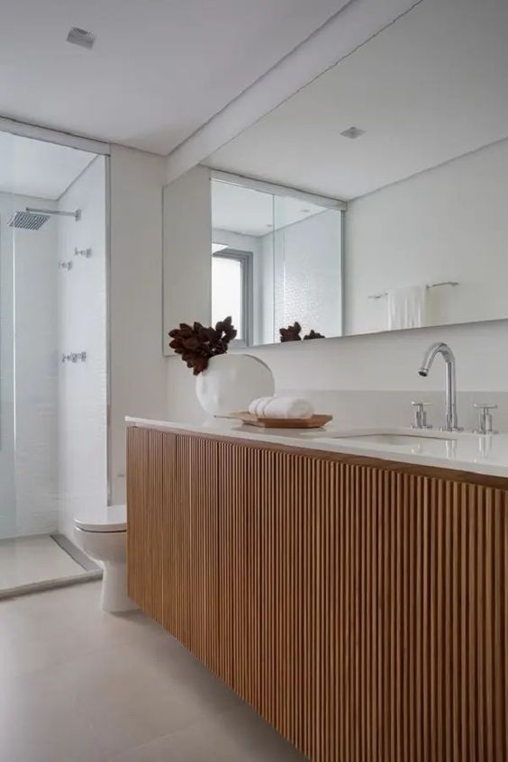 an elegant and chic stained fluted vanity with a white stone countertop, a vase with foliage and a large mirror over it