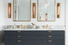 an elegant bathroom clad with white marble, with a black fluted vanity, gold touches, gold frame mirrors and sconces