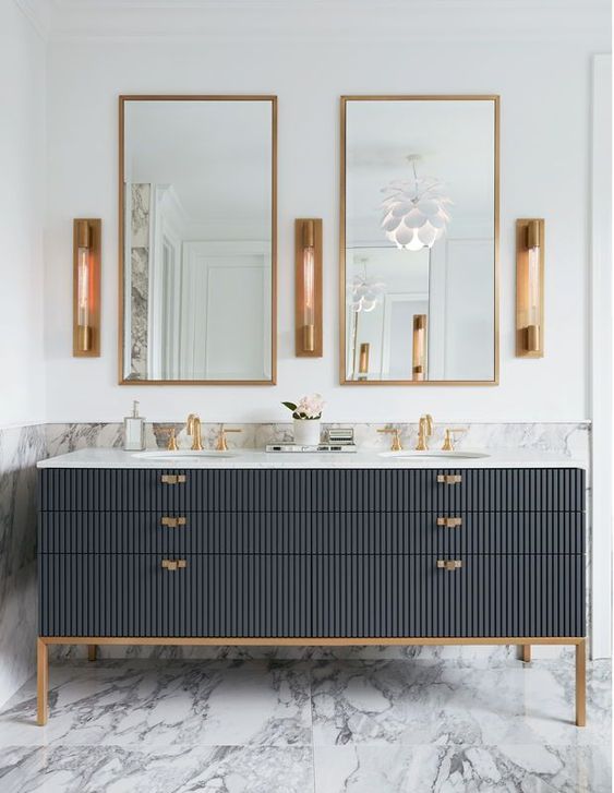 an elegant bathroom clad with white marble, with a black fluted vanity, gold touches, gold frame mirrors and sconces
