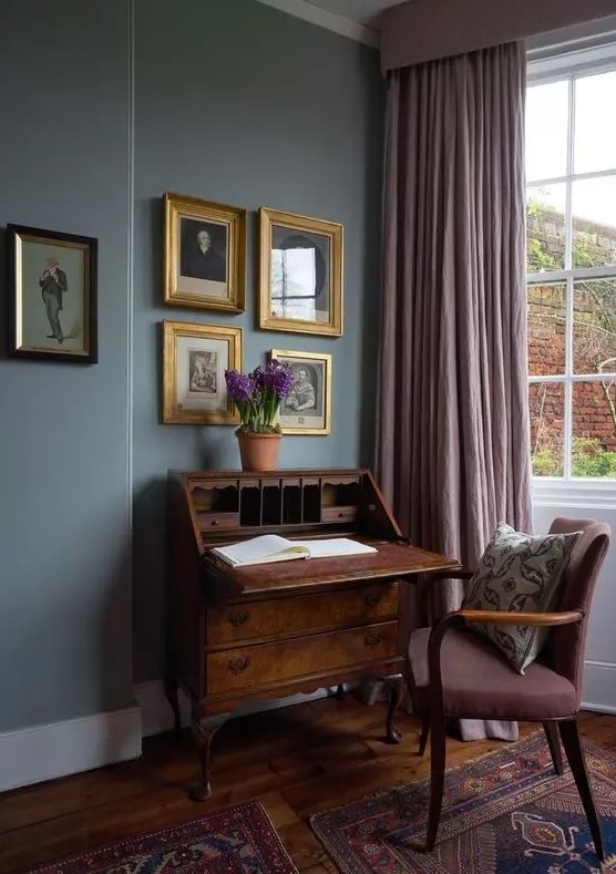 An exquisite vintage working nook with grey walls, a vintage dark stained bureau and a mauve chair, mauve curtains and a gallery wall