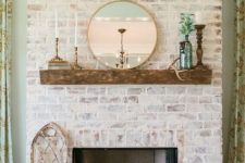 a beautiful whitewashed red brick fireplace with a rough wooden mantel, candles, greenery and a round mirror