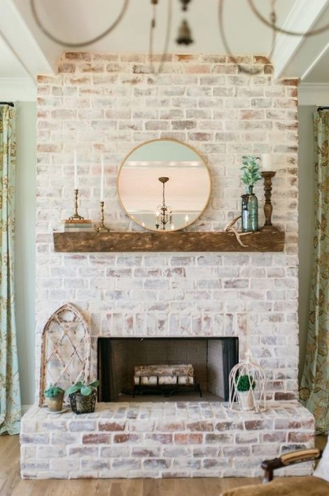 a beautiful whitewashed red brick fireplace with a rough wooden mantel, candles, greenery and a round mirror