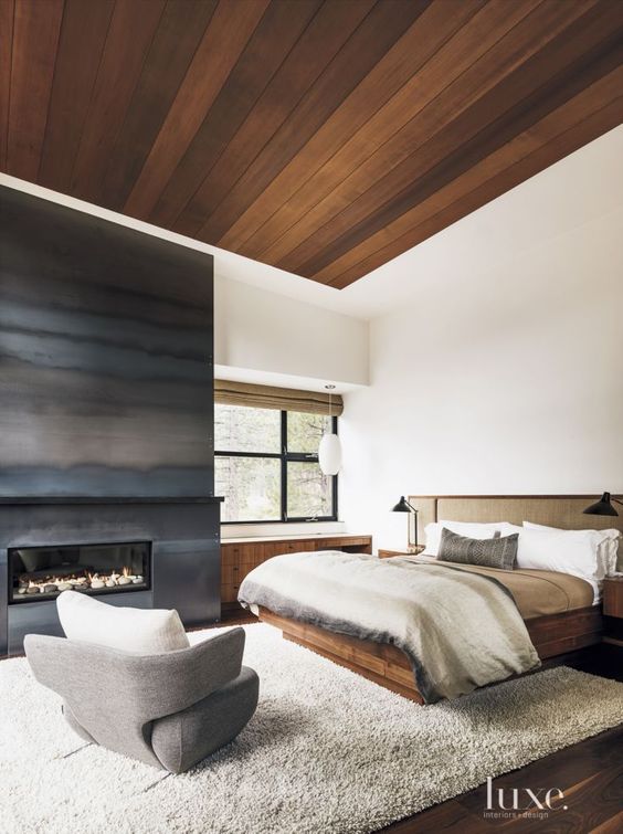 a bedroom with a stained wood ceiling, a metal fireplace, stained furniture, neutral textiles is welcoming