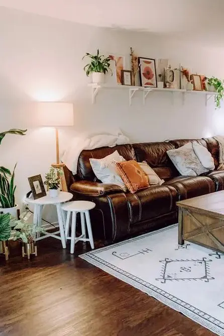 a boho living room with a brown leather sofa, a wooden coffee table, side tables, a shelf with art and lots of greenery