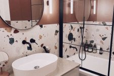 a bright bathroom with rust walls, bold terrazzo, a bathtub with a screen, a vanity, a sink, a round mirror and black fixtures
