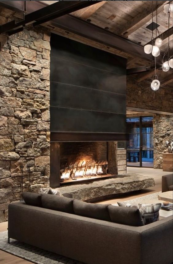 a chalet living room with stone walls, a fireplace done with metal, taupe seating furniture and pendant lamps