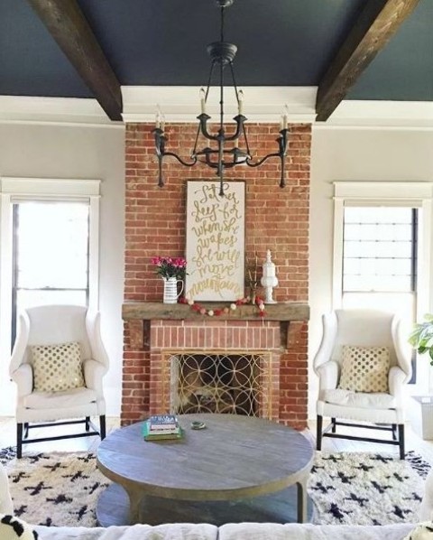 a chic living room with a red brick fireplace, a black ceiling with dark stained beams, white furniture and a round table