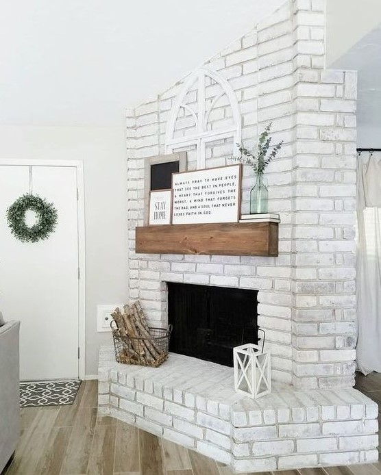 a chic whitewashed brick fireplace with a wooden mantel, artworks and eucalyptus, a wire basket with firewood