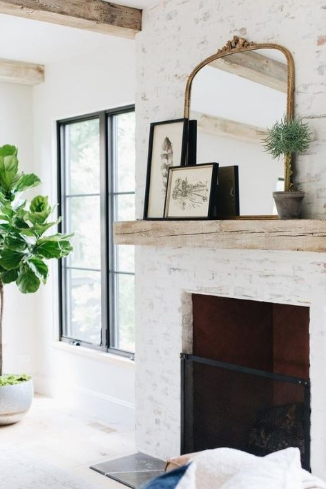 a chic whitewashed fireplace of brick and with a metal screen, a wooden mantel with artworks and a chic mirror