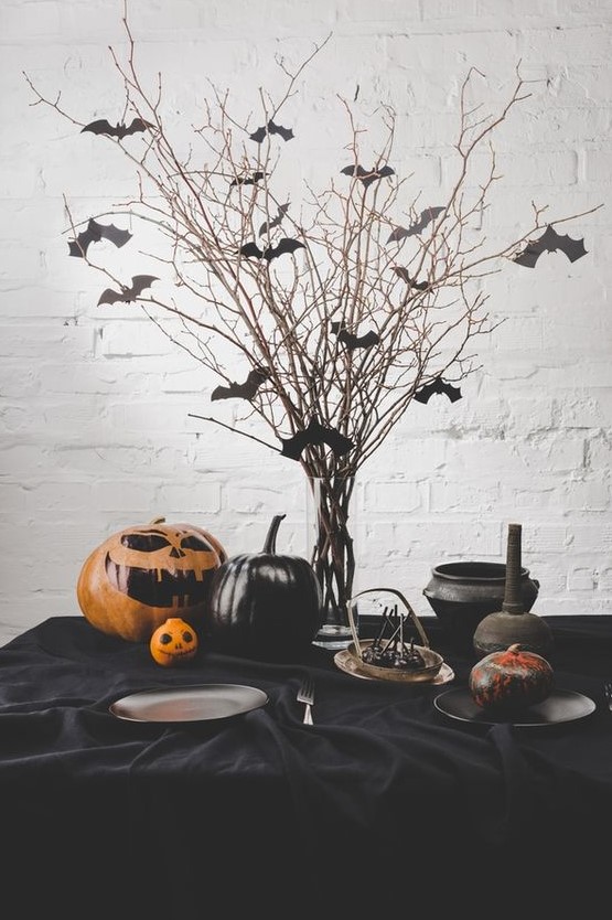 a classic Halloween table setting with bats on branches, black pumpkins and a painted orange one, a black tablecloth