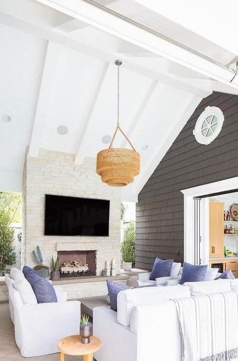 a coastal outdoor living room with a white brick fireplace, white furniture, blue pillows, a woven pendant lamp and potted plants