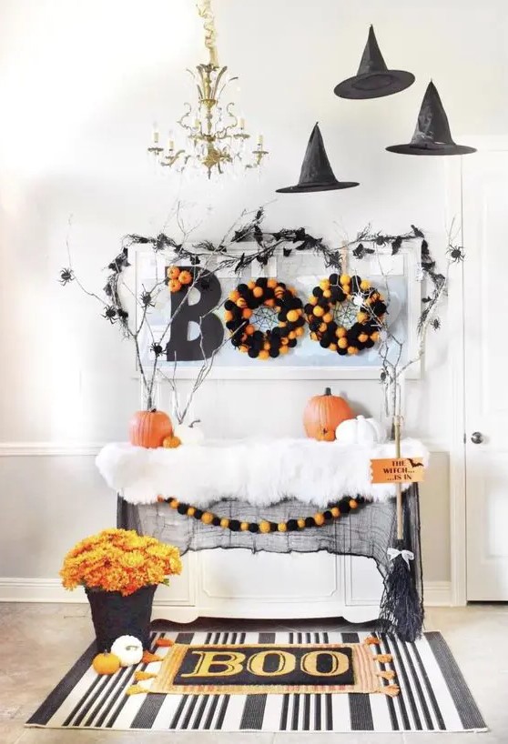 a console table styled for Halloween in a bold and fun way, with black witches' hats, faux fur, pumpkins, wreaths and bold blooms, branches and spiders