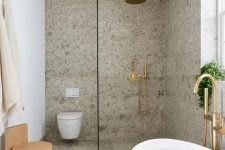 a cool modern bathroom clad with grey terrazzo, with a shower space, an oval tub, a wooden stool and some gold and brass fixtures