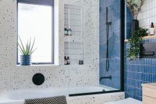 a cool modern bathroom with white terrazzo, a bold blue skinny tile wall, a tub with a window over it, a stool and some greenery
