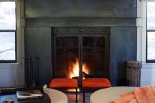 a cozy fireplace nook with a fireplace clad with metal, a duo of orange stools, round ottomans and a reclaimed wood ceiling
