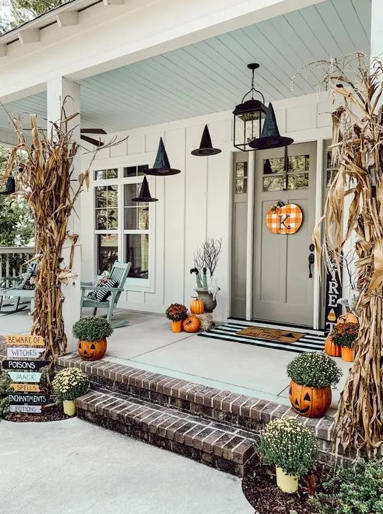 a cozy rustic Halloween porch with dried corn husks, pumpkins, pumpkin planters with blooms and witches' hats