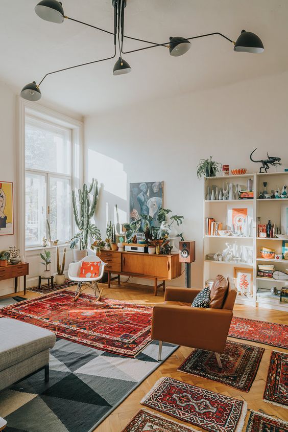 A mid century boho living room with a grey sofa, printed rugs, a stained credena, an amber chair, potted plants and artwork