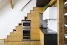 a modern kitchen with stained cabinets built into the stairs, black countertops, upper white cabinets and greenery