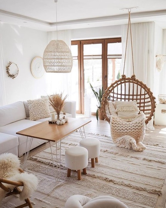 a neutral boho living room with white furniture, a suspended chair, a wooden table and stools