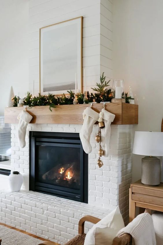 a pretty white brick fireplace with a mantel styled for Christmas, with stockings, bells, evergreens and lights