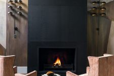 a refined fireplace nook with a fireplace clad with black metal, striped chairs, a tiny side table and cool wall lamps