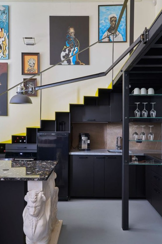 A staircase with a built in black kitchen, open glass shelves, a kitchen island, some decor and a black sconce