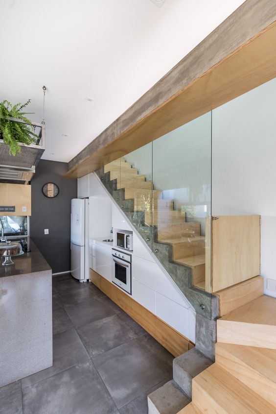 a stylish modern kitchen with sleek white cabinets built in under the stairs, a concrete kitchen island and greenery
