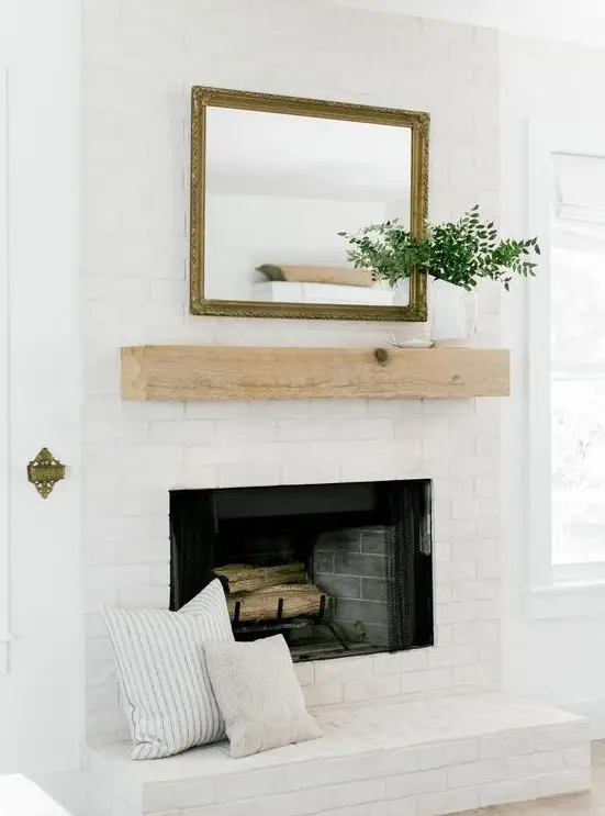 a stylish whitewashed brick fireplace with a wooden mantel, a mirror and greenery plus printed pillows for a cozy feel