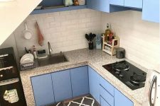 a tiny blue kitchen built under the staircase, with a subway tile backsplash and terrazzo countertops and black rugs