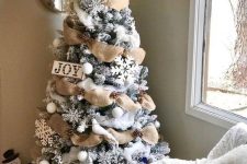 a flocked farmhouse Christmas tree with burlap ribbon, pinecones, snowflakes and a sign plus some snowy branches on top