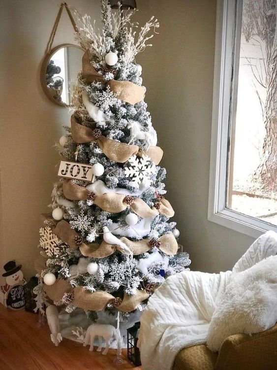 a flocked farmhouse Christmas tree with burlap ribbon, pinecones, snowflakes and a sign plus some snowy branches on top