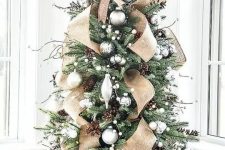 a gorgeous tabletop Christmas tree with silver and metallic ornaments, pinecones, twigs and a large burlap bow on top
