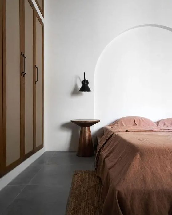 A minimal earthy bedroom with a built in wardrobe, a bed with rust bedding, a metal nightstand and a black sconce