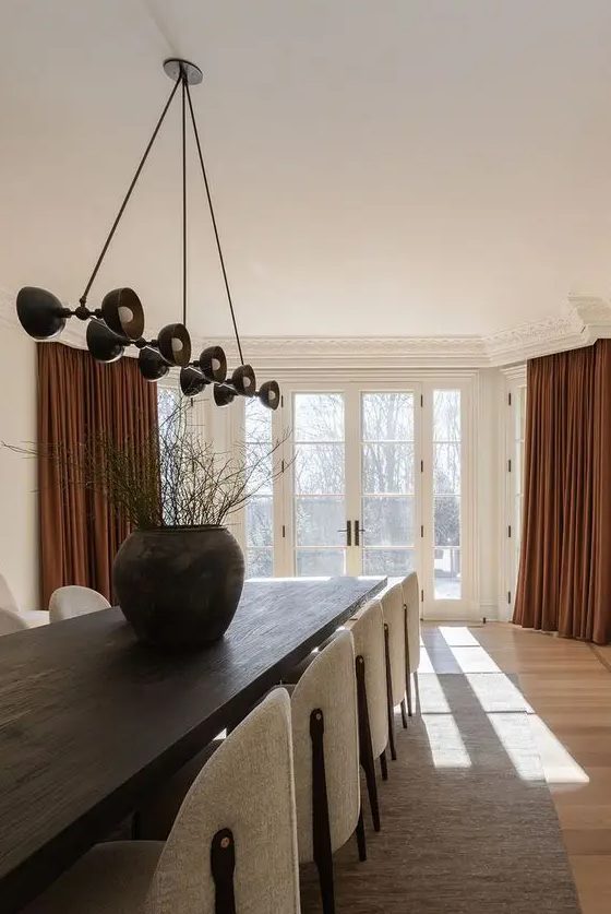 a refined minimalist dining space with a dark-stained table, neutral curved chairs, a black pendant lamp and rust-colored curtains