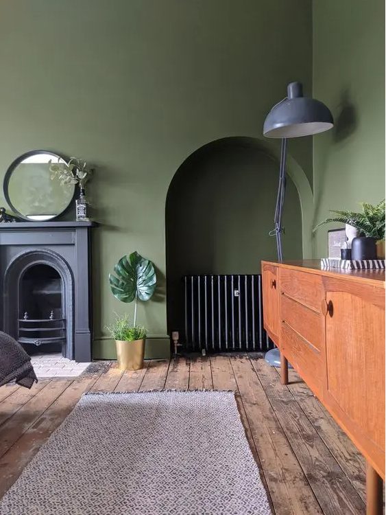 A vintage olive green living room with a non working fireplace, a niche with a credenza, a stained sideboard, a grey floor lamp and a mirror