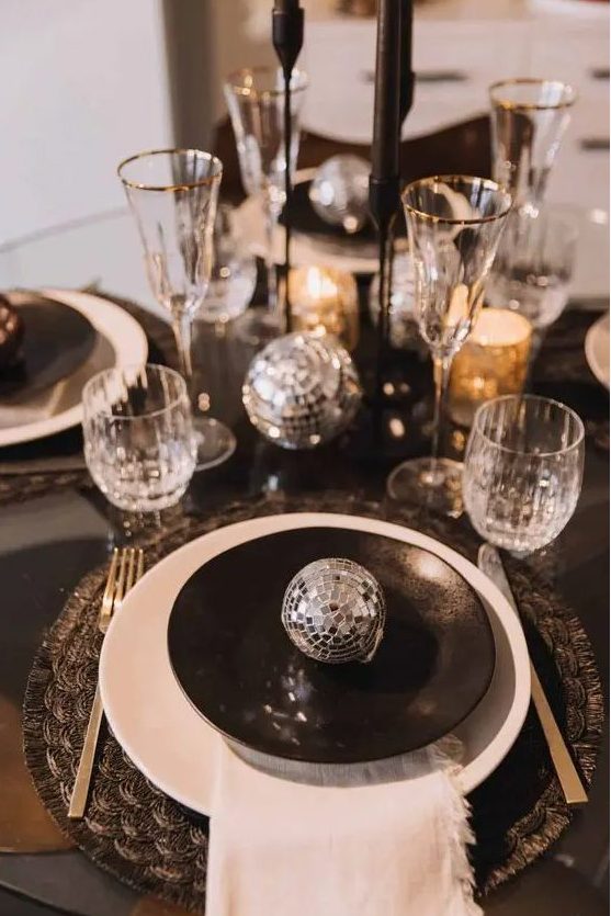 a black and silver NYE tablescape with black printed placemats, black and white plates, black candles and silver disco balls