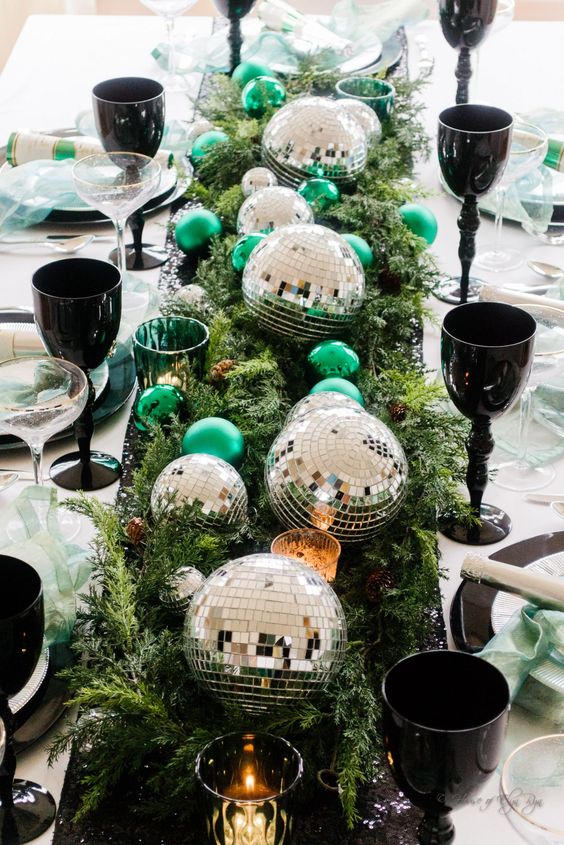 a bright NYE party table with evergreens, silver disco balls, green ornaments and black glasses plus green napkins