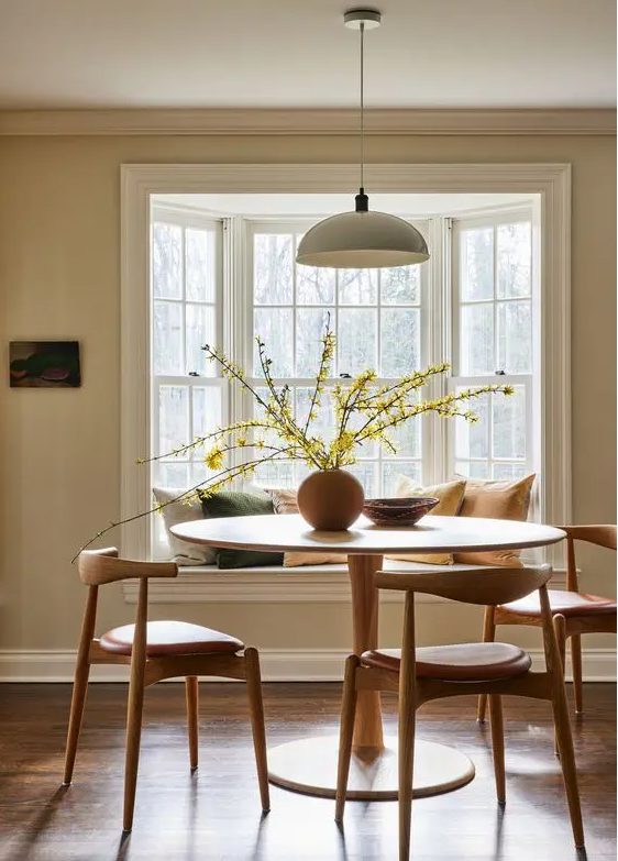 An earthy dining room with a bay window with a window seat, a round table and mid century modern chairs, a pendant lamp