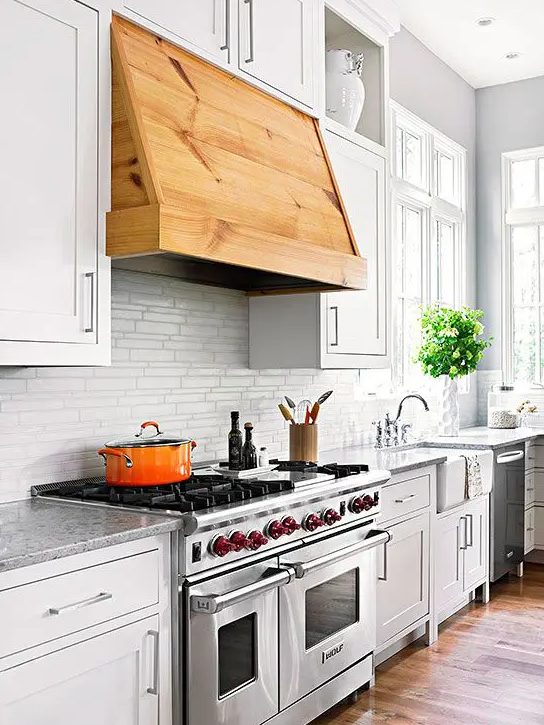 a white farmhouse kitchen with a white tile backsplash and grey countertops, a stained hood that adds a warm touch to the space