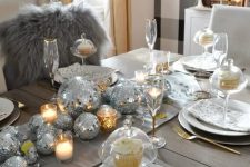 23 a shiny holiday tablescape with silver disco balls forming a table runner, candles, white porcelain and gold cutlery